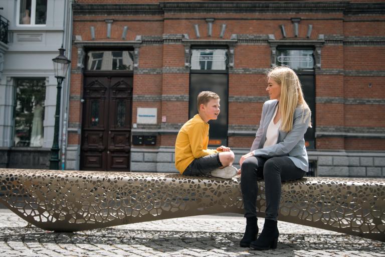 Blond jongetje met blonde dame in gesprek op metalen bank in stad