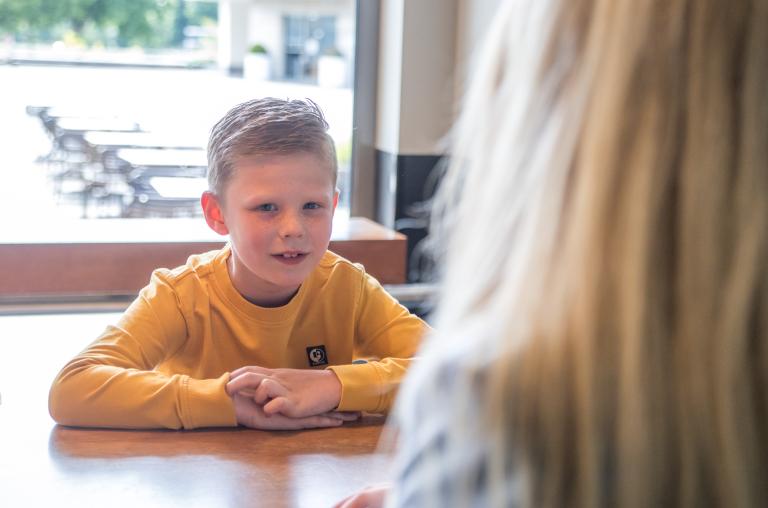 Blond jongetje luistert naar dame aan hoge tafel